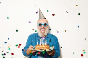 Portrait of excited senior man in sunglasses smiling at camera while standing under the confetti and holding cake